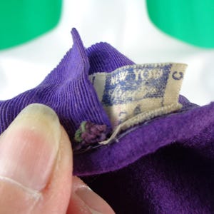 Peut inclure: Un v&ecirc;tement en tissu violet profond dot&eacute; d'un col c&ocirc;tel&eacute; et d'une &eacute;tiquette int&eacute;rieure rectangulaire blanche. L'&eacute;tiquette affiche le texte New York et la lettre C &agrave; l'encre bleue. Le tissu pr&eacute;sente une texture douce semblable &agrave; du feutre.