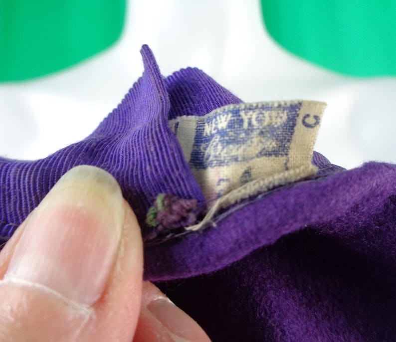 Peut inclure: Un v&ecirc;tement en tissu violet profond dot&eacute; d'un col c&ocirc;tel&eacute; et d'une &eacute;tiquette int&eacute;rieure rectangulaire blanche. L'&eacute;tiquette affiche le texte New York et la lettre C &agrave; l'encre bleue. Le tissu pr&eacute;sente une texture douce semblable &agrave; du feutre.