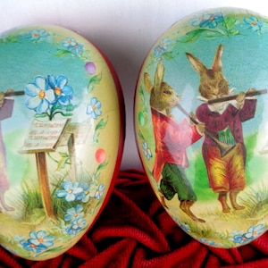 May include: Two vintage Easter egg-shaped candy containers with a pastel blue background. Each container features a pair of rabbits playing musical instruments. The rabbits are dressed in red, yellow, and blue clothing. The containers are decorated with blue flowers and green leaves.