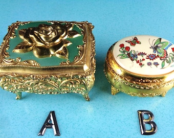 May include: Two metal trinket boxes labeled A and B. Box A is a rectangular gold-tone container with a green lid featuring a raised gold rose. Box B is a round gold-tone container with a white lid featuring a colorful butterfly and floral print.