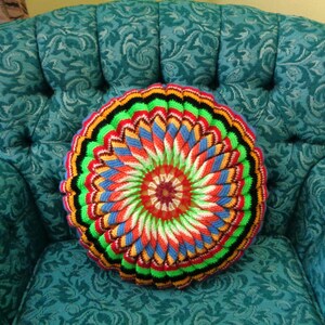 May include: A circular, multi-coloured crochet cushion with a floral pattern sits on a teal armchair. The cushion's design includes rings of red, orange, blue, green, and black, offering a vivid contrast.