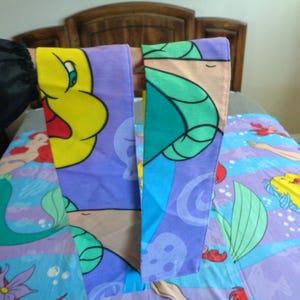 May include: A bed set featuring characters from The Little Mermaid. The set includes a pillowcase with a yellow character and a blue and purple comforter with Ariel and Flounder. The bedding has a cartoon style.