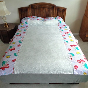 May include: A bed with a wooden headboard is covered with a light blue and white patterned bedspread featuring red-haired mermaids and other sea creatures. A white sheet covers the center of the bed.