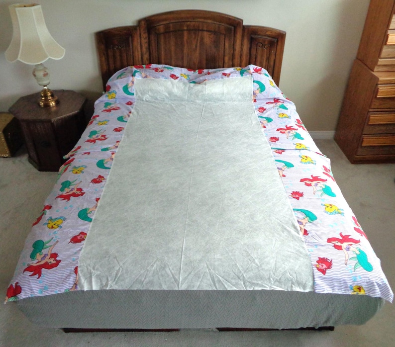 May include: A bed with a wooden headboard is covered with a light blue and white patterned bedspread featuring red-haired mermaids and other sea creatures. A white sheet covers the center of the bed.