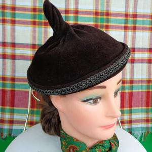 Può includere: Un cappello di velluto marrone con un bordo intrecciato nero e una cima a punta. Il cappello è su una testa di manichino con una sciarpa verde e marrone.