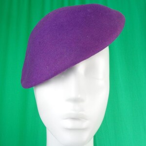 Peut inclure: Un b&eacute;ret violet uni avec une texture feutr&eacute;e lisse. Le chapeau est positionn&eacute; l&eacute;g&egrave;rement de biais sur une t&ecirc;te de mannequin blanche, mettant en valeur sa forme arrondie classique et sa couleur violette vive sur toute la surface du couvre-chef.