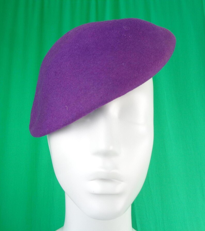 Peut inclure: Un b&eacute;ret violet uni avec une texture feutr&eacute;e lisse. Le chapeau est positionn&eacute; l&eacute;g&egrave;rement de biais sur une t&ecirc;te de mannequin blanche, mettant en valeur sa forme arrondie classique et sa couleur violette vive sur toute la surface du couvre-chef.