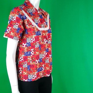 Op de afbeelding: Een rood patchwork bloesje met korte mouwen en witte kanten afwerking. Het bloesje, met knopen aan de voorkant, wordt getoond op een mannequin. Het patroon bevat rode, blauwe en witte bloemen.