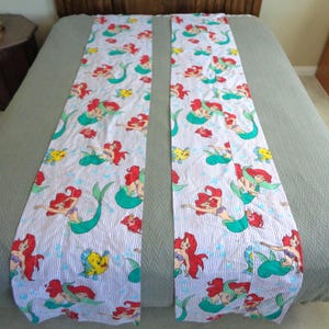 May include: Two long, rectangular fabric pieces featuring a repeating pattern of a mermaid with red hair and a green tail, along with fish and other sea creatures. The background has blue and white vertical stripes.