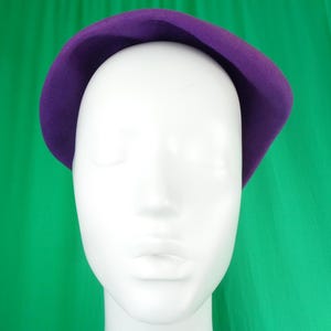 Peut inclure: Un b&eacute;ret en feutre violet uni avec une calotte arrondie et lisse et un bord l&eacute;g&egrave;rement incurv&eacute;. Le chapeau est pr&eacute;sent&eacute; sur une t&ecirc;te de mannequin blanche unie, mettant en valeur la couleur violette profonde et vibrante ainsi que la texture douce.