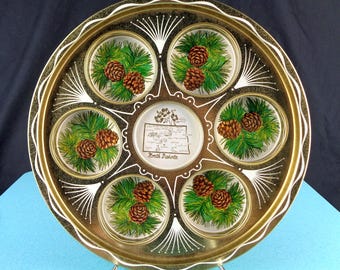 May include: A round gold-toned metal serving tray featuring five circular indentations decorated with green pine needles and brown pinecones. The center displays a map of North Dakota with text labels for various cities and the state name.