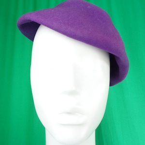 Peut inclure: Un b&eacute;ret en feutre violet uni avec une calotte arrondie et lisse et un bord l&eacute;g&egrave;rement incurv&eacute;, pos&eacute; de biais sur une t&ecirc;te de mannequin blanche. Le chapeau pr&eacute;sente une texture uniforme et une couleur violette vive sur toute sa surface.