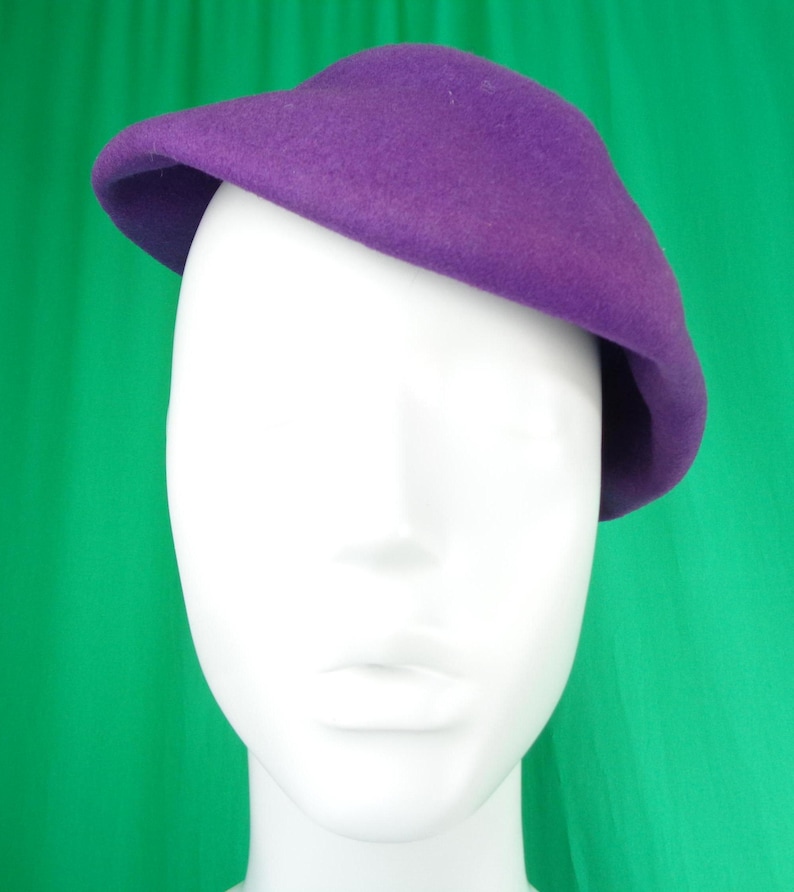 Peut inclure: Un b&eacute;ret en feutre violet uni avec une calotte arrondie et lisse et un bord l&eacute;g&egrave;rement incurv&eacute;, pos&eacute; de biais sur une t&ecirc;te de mannequin blanche. Le chapeau pr&eacute;sente une texture uniforme et une couleur violette vive sur toute sa surface.