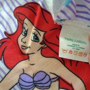 May include: Ariel the Little Mermaid cartoon character on a striped background. The character has red hair, blue eyes, and a purple seashell bra. The tag says "TWIN/JUMEAU" and "MADE IN FABRIQUE AU CANADA".