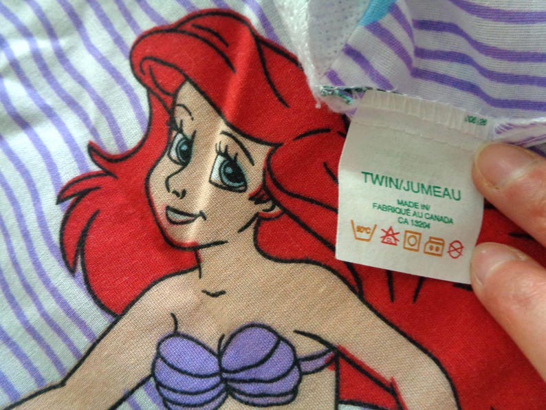 May include: Ariel the Little Mermaid cartoon character on a striped background. The character has red hair, blue eyes, and a purple seashell bra. The tag says "TWIN/JUMEAU" and "MADE IN FABRIQUE AU CANADA".