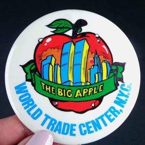 May include: A white button with a red apple design, featuring the Twin Towers in blue and yellow. The green banner reads "THE BIG APPLE" and "WORLD TRADE CENTER, N.Y.C." in blue text, a vintage souvenir.