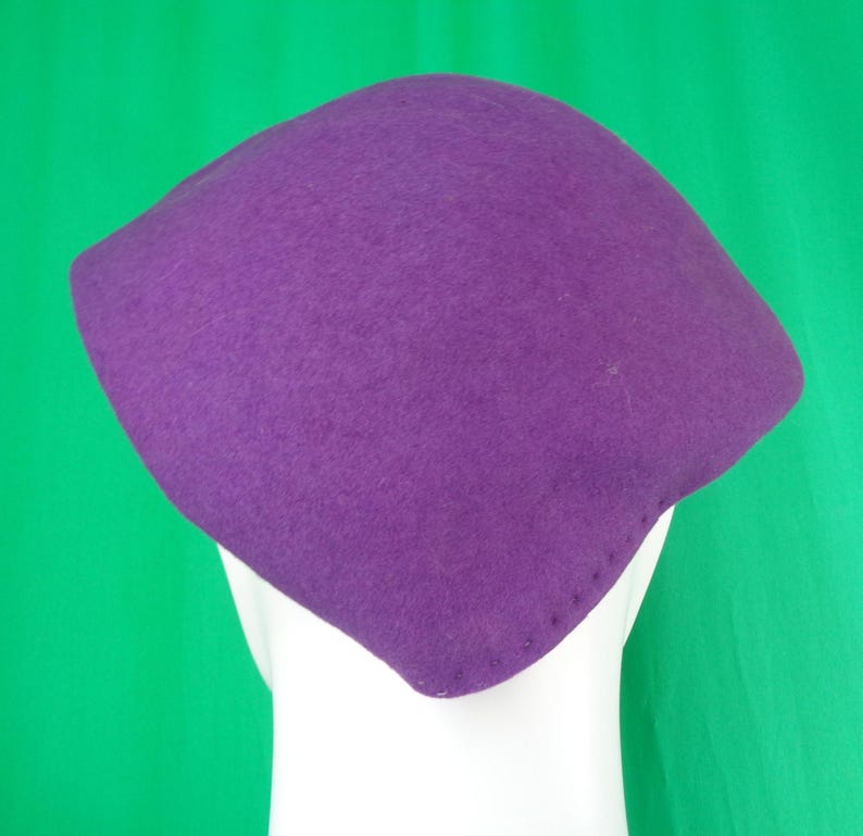 Peut inclure: Un chapeau en feutre violet uni avec une forme structur&eacute;e, angulaire et &agrave; quatre coins. Le chapeau pr&eacute;sente une texture lisse et mate ainsi qu'une s&eacute;rie de petites coutures r&eacute;guli&egrave;rement espac&eacute;es le long du bord inf&eacute;rieur.