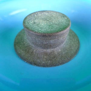 May include: A close-up of a gray bottle stopper with a circular top and a wide, flat base. The stopper is set against a bright turquoise background, creating a striking contrast. The stopper appears to be made of a textured material.
