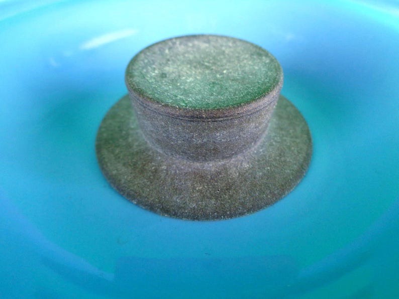 May include: A close-up of a gray bottle stopper with a circular top and a wide, flat base. The stopper is set against a bright turquoise background, creating a striking contrast. The stopper appears to be made of a textured material.