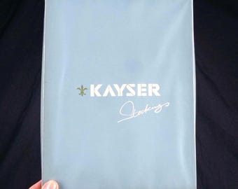 May include: A rectangular light blue package with white text that reads KAYSER and Stockings in a script font. A small gold fleur-de-lis symbol is positioned to the left of the word KAYSER. The packaging has a smooth, matte finish.