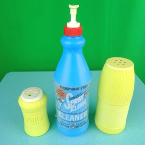 May include: A blue Spray Kleen cleaner bottle with a red cap and white pump, flanked by two yellow containers. The bottle has a label with text. The containers are yellow with white tops. The background is green.