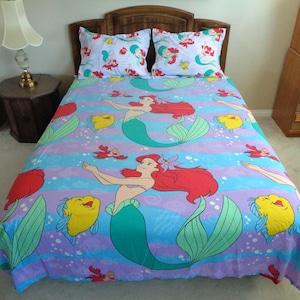 May include: A bed set featuring a mermaid theme. The bedding includes a comforter and two pillowcases with a repeating pattern of a mermaid with red hair, yellow fish, and red crabs. The background has blue and purple stripes.