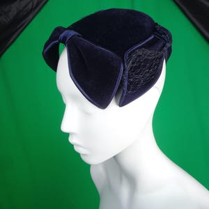 May include: A navy blue velvet hat with a large bow detail. The hat features a net veil and is displayed on a white mannequin head. The hat is a vintage style, suitable for special occasions.