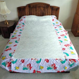 May include: A bed with a wooden headboard is covered with a light blue and white striped sheet. The sheet has a colorful pattern of mermaids, fish, and other sea creatures. A white mattress pad covers the center of the bed.