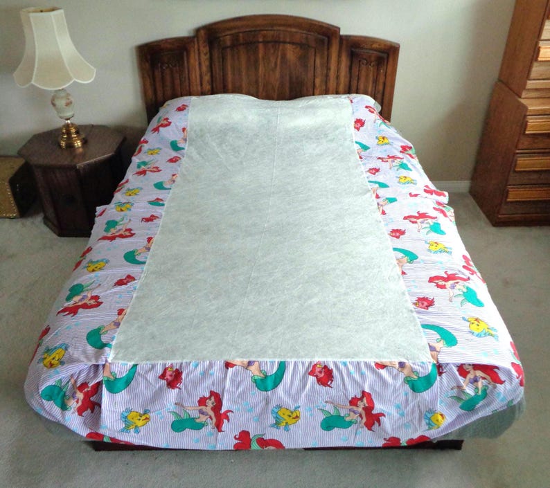 May include: A bed with a wooden headboard is covered with a light blue and white striped sheet. The sheet has a colorful pattern of mermaids, fish, and other sea creatures. A white mattress pad covers the center of the bed.