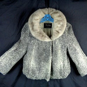 May include: A vintage grey fur coat with a light-coloured fur collar. The coat is hanging on a blue hanger against a dark blue background. The coat has a textured pattern and a label that reads "N. Chen".