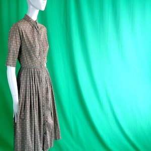 May include: A vintage button-down dress in a brown and teal geometric pattern. The dress features short sleeves, a collared neckline, and a pleated skirt. The dress is displayed on a mannequin against a green backdrop.