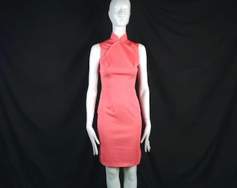 May include: A sleeveless coral-colored sheath dress featuring a high mandarin collar and an asymmetrical crossover neckline. The garment is fitted through the torso and waist, falling to a straight hemline above the knee, displayed on a mannequin.