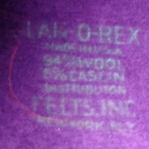 Peut inclure: Une surface en tissu violet avec du texte blanc imprim&eacute; indiquant LAM-O-REX, MADE IN U.S.A., 94% WOOL, 6% CASEIN, DISTRIBUTOR, FELTS, INC. Le mat&eacute;riau pr&eacute;sente une texture feutr&eacute;e douce avec des lettres blanches clairement visibles sur le fond violet.