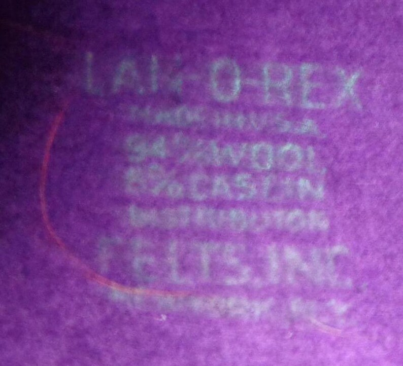 Peut inclure: Une surface en tissu violet avec du texte blanc imprim&eacute; indiquant LAM-O-REX, MADE IN U.S.A., 94% WOOL, 6% CASEIN, DISTRIBUTOR, FELTS, INC. Le mat&eacute;riau pr&eacute;sente une texture feutr&eacute;e douce avec des lettres blanches clairement visibles sur le fond violet.