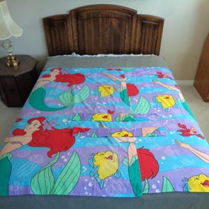 May include: A colorful bedspread featuring a mermaid theme. The design includes mermaids with red hair, yellow fish, and red crabs on a blue and purple striped background. The bedspread is on a bed with a wooden headboard.