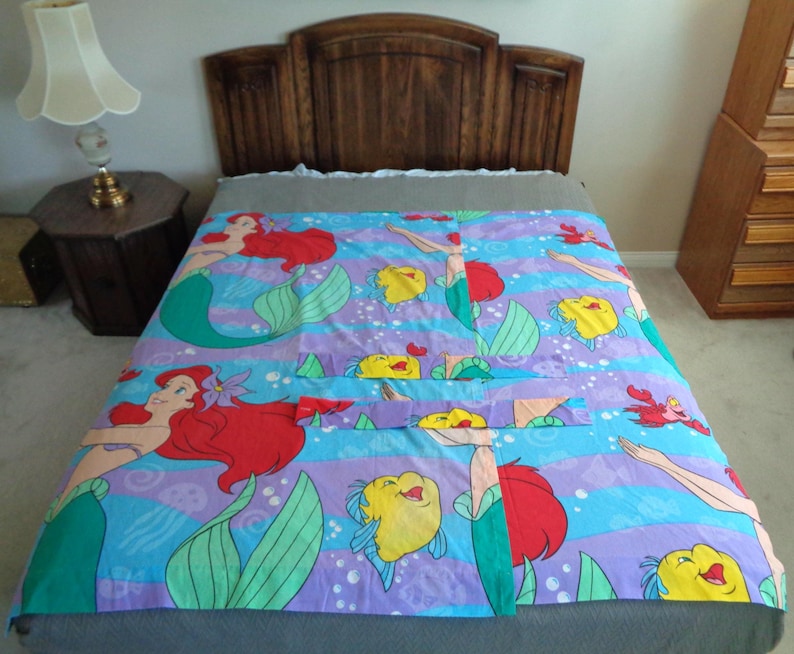 May include: A colorful bedspread featuring a mermaid theme. The design includes mermaids with red hair, yellow fish, and red crabs on a blue and purple striped background. The bedspread is on a bed with a wooden headboard.