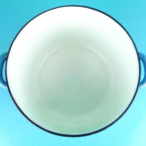 May include: A white enamel cast iron Dutch oven with blue handles and a black rim. The interior is white, and the pot is empty. The handles are curved and attached to the sides of the pot. The background is a solid light blue.