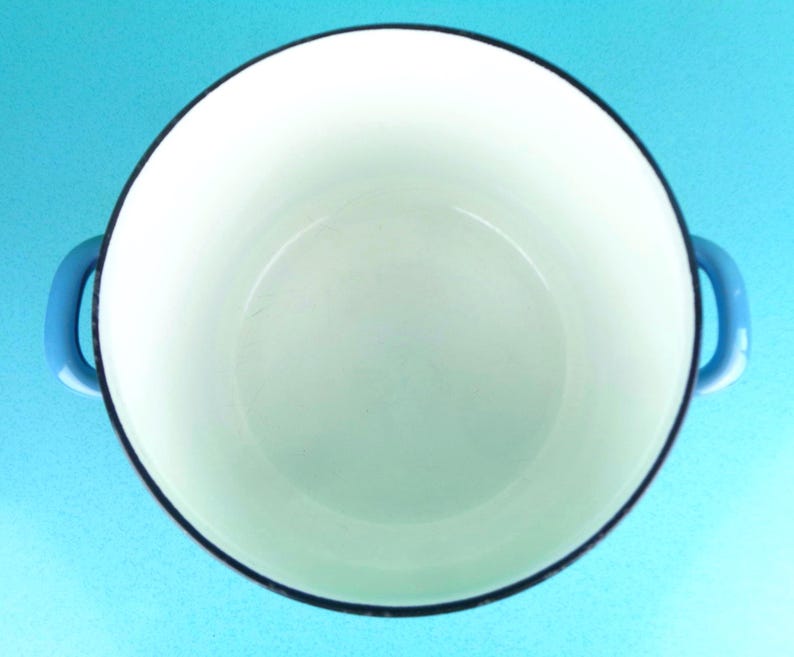 May include: A white enamel cast iron Dutch oven with blue handles and a black rim. The interior is white, and the pot is empty. The handles are curved and attached to the sides of the pot. The background is a solid light blue.