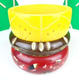 May include: Three stacked vintage bangles. The top bangle is bright yellow with carved floral designs. The middle bangle is dark brown with gold-coloured leaf accents. The bottom bangle is red with white and black flecks.