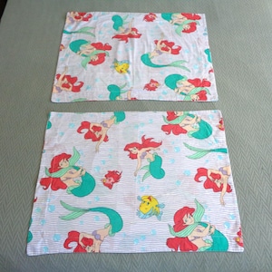 May include: Two white pillowcases with a repeating pattern of a red-haired mermaid, teal tail, and yellow fish. The design is set against a light blue striped background with small blue bubbles. The pillowcases are likely for a child's bed.