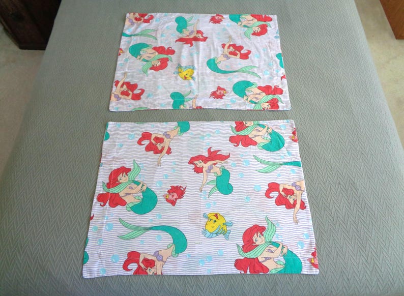 May include: Two white pillowcases with a repeating pattern of a red-haired mermaid, teal tail, and yellow fish. The design is set against a light blue striped background with small blue bubbles. The pillowcases are likely for a child's bed.