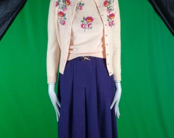 May include: A pale peach cardigan and matching short-sleeve sweater set featuring embroidered pink and yellow floral motifs with small bead accents. The set is paired with a solid dark purple pleated skirt featuring a gold-tone metal belt buckle.