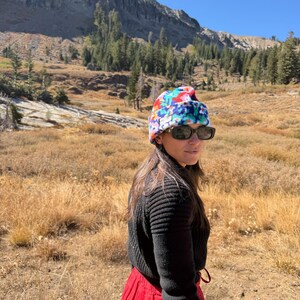 May include: A beanie with a multi-colored floral pattern in shades of red, blue, and green, worn with black sunglasses, a black ribbed long-sleeve sweater, and a solid red skirt, set against a backdrop of a mountain landscape.