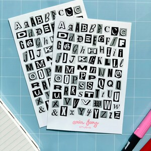May include: Two white sticker sheets with black and gray cut-out style letters. The sheets are arranged on a light blue grid surface. The text "erin fong STUDIO" is printed in pink. A notebook and two markers are also visible.