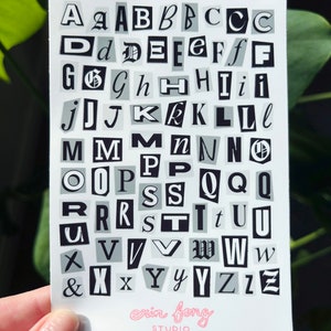 May include: A white card featuring a collage of black and gray letters in various fonts. The letters are cut out, resembling a ransom note style. The artist's name, "erin fong STUDIO," is printed in pink at the bottom.