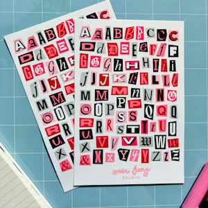 May include: Two white art prints with a collage of letters in pink, red, black, and gray. The prints feature the text "erin fong STUDIO" in pink. The background is a light blue grid.