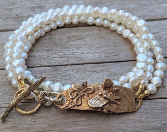 May include: A multi-strand pearl bracelet with a gold clasp featuring a floral design. The bracelet is made of white pearls and has a gold toggle clasp.