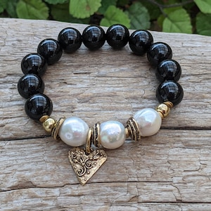 May include: Black onyx and white pearl bracelet with a gold heart charm. The bracelet is made with round beads and has a simple design.
