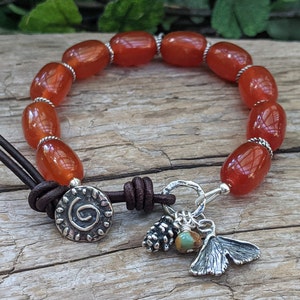 Op de afbeelding: Een handgemaakte armband met ovale, oranje-rode kralen en zilverkleurige afstandhouders. De armband heeft een bruin leren koordsluiting en decoratieve bedels, waaronder een dennenappel en een ginkgoblad.