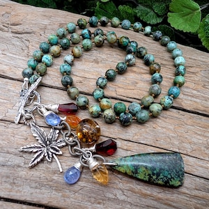 Puede incluir: Un collar de cuentas con piedras redondas de color turquesa con vetas oscuras. Un cierre de barra plateado sostiene un grupo de dijes, incluyendo una hoja de plata, un gran colgante triangular de turquesa y varias cuentas de cristal facetado.
